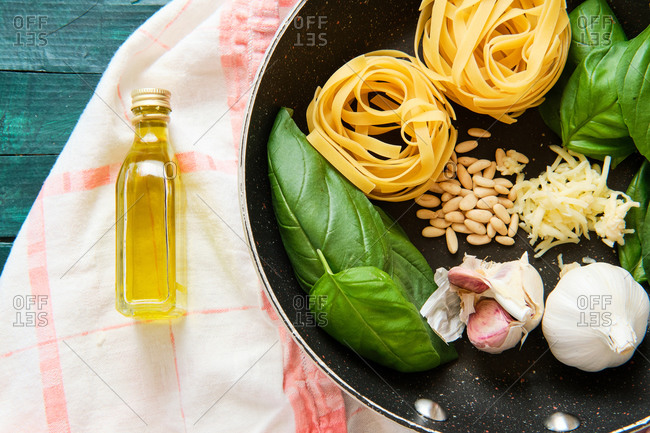Top view of bottle of olive oil near frying pan with raw tagliatelle rolls surrounded by heads of garlic with green basil leaves and crispy pine nuts