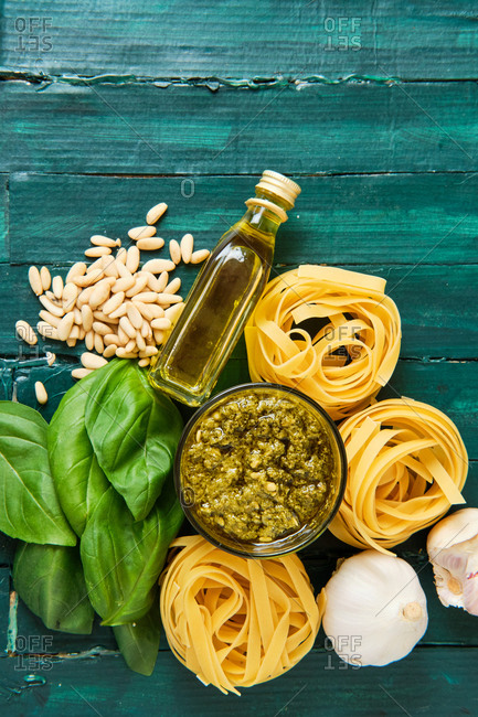 Top view of composition of dry pasta rolls near bowl with pesto sauce and fresh basil leaves with crushed garlic and pine nuts near bottle with olive oil