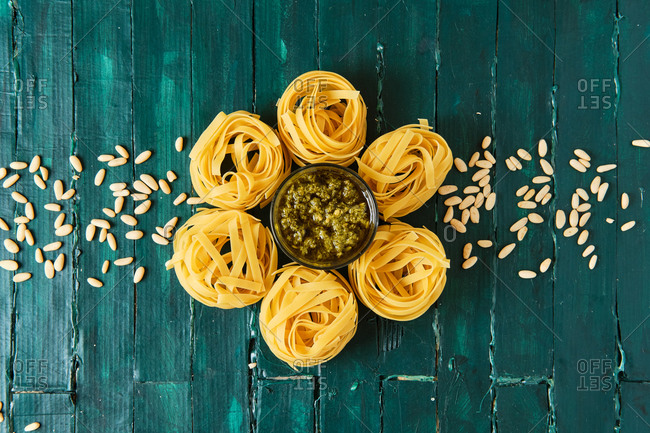 From above of tasty pesto salsa with olive oil surrounded by uncooked pasta rolls and spilled crunchy pine nuts on wooden table