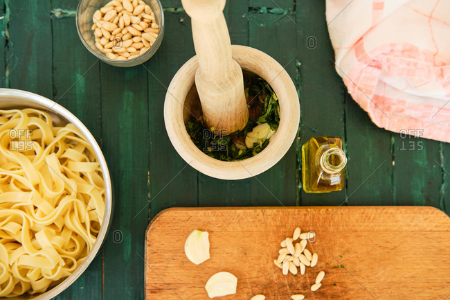 Top view of bowl with freshly cooked Italian pasta near wooden pounder with pestle and partially pounded basil with garlic gloves and pine nuts near bottle with olive oil
