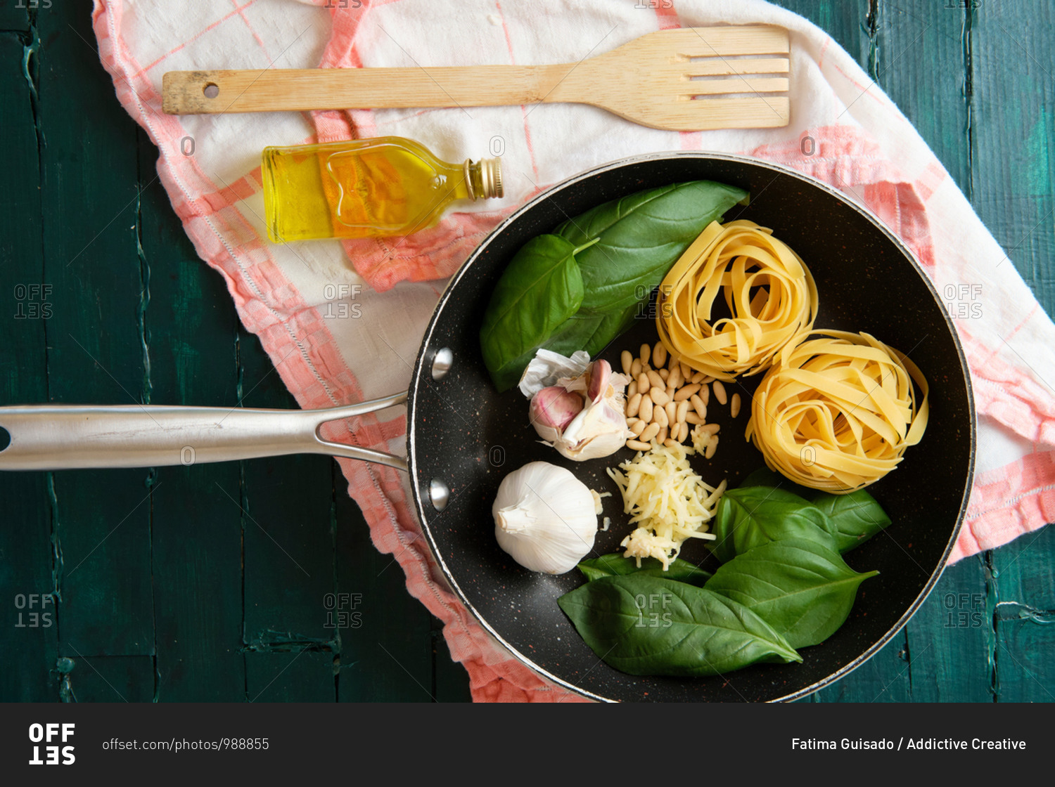 Top view of frying pen with uncooked pasta rolls near fresh basil leaves and heads of garlic with pine nuts and bottle of olive oil near wooden spatula on towel