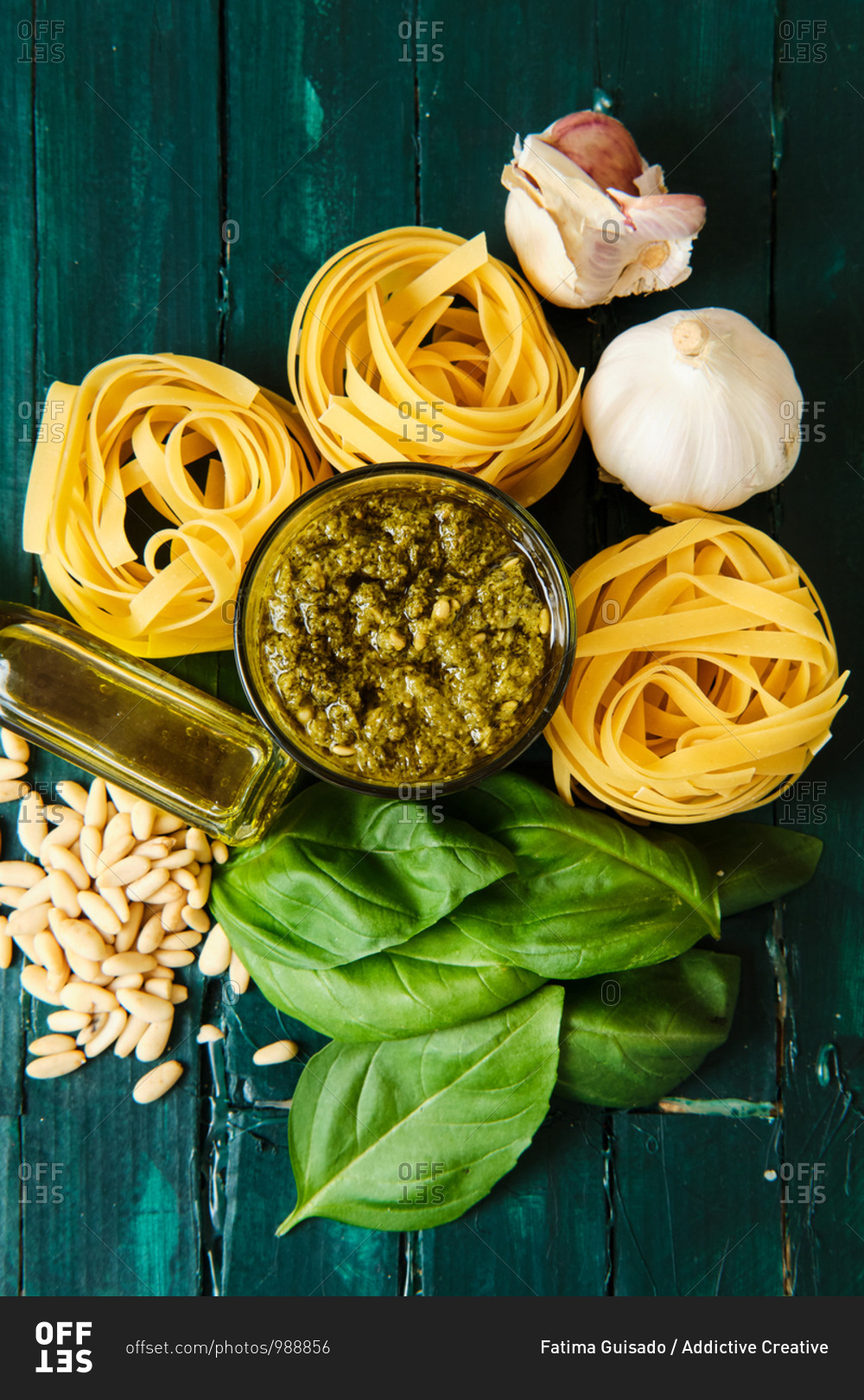 Top view of composition of dry pasta rolls near bowl with pesto sauce and fresh basil leaves with crushed garlic and pine nuts near bottle with olive oil