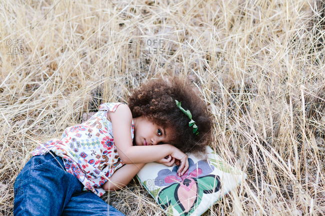Adorable calm mulatto girl in trendy dress lying on plaid on wheat field on summer day and looking at camera