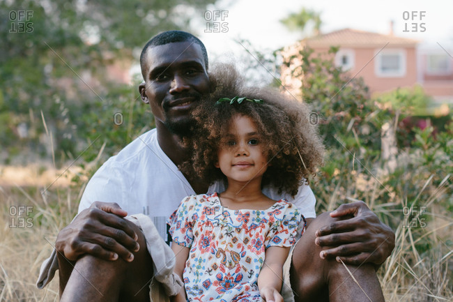 Smiling African American father sitting on meadow with adorable glad daughter between legs and looking at camera happily in countryside area during summer day