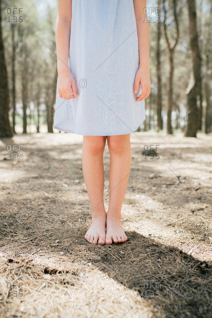 Crop unrecognizable slender barefoot child in pale blue dress standing on dry terrain in alley on sunny day in countryside