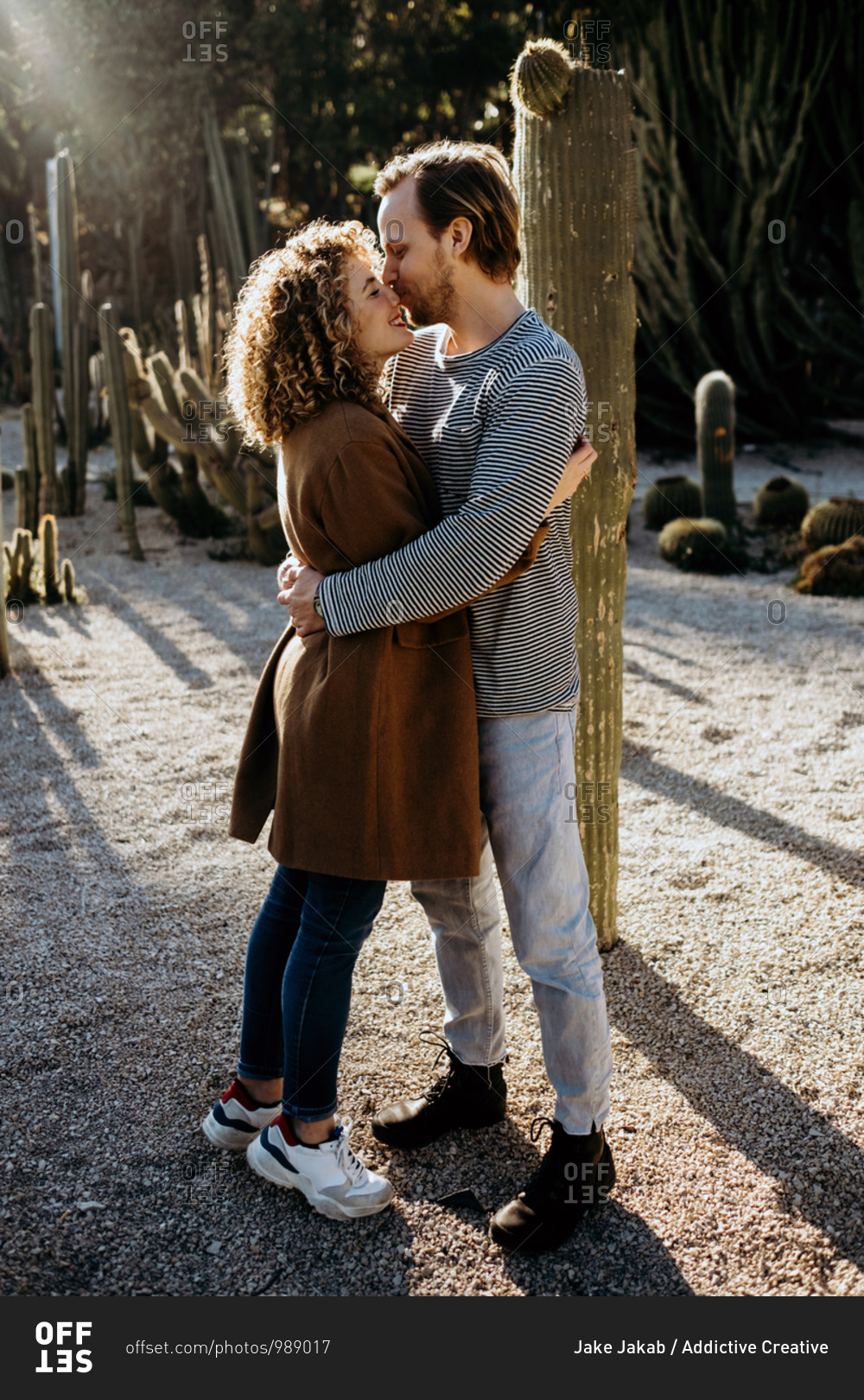 Side view of cheerful man and woman kissing and embracing each other while spending summer day on blurred background of cactuses in park in Barcelona, Spain