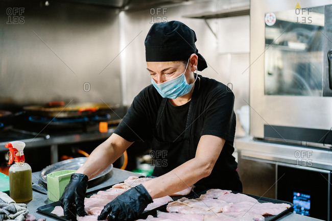 Serious female cook wearing apron respirator and latex gloves preparing fresh meat on trays while working in modern restaurant kitchen