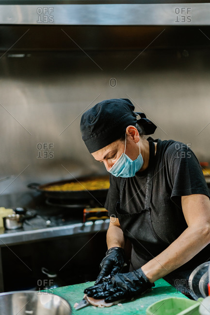 Serious female cook wearing apron respirator and latex gloves cutting fresh meat on cutting board while working in modern restaurant kitchen