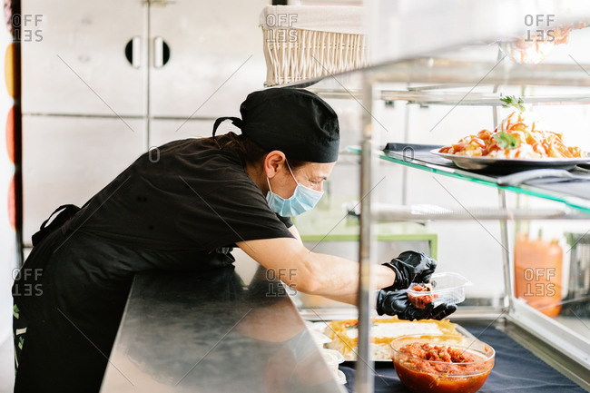 Side view focused waitress wearing face mask and gloves putting delicious dish into takeaway plastic container while working in cafe during coronavirus pandemic