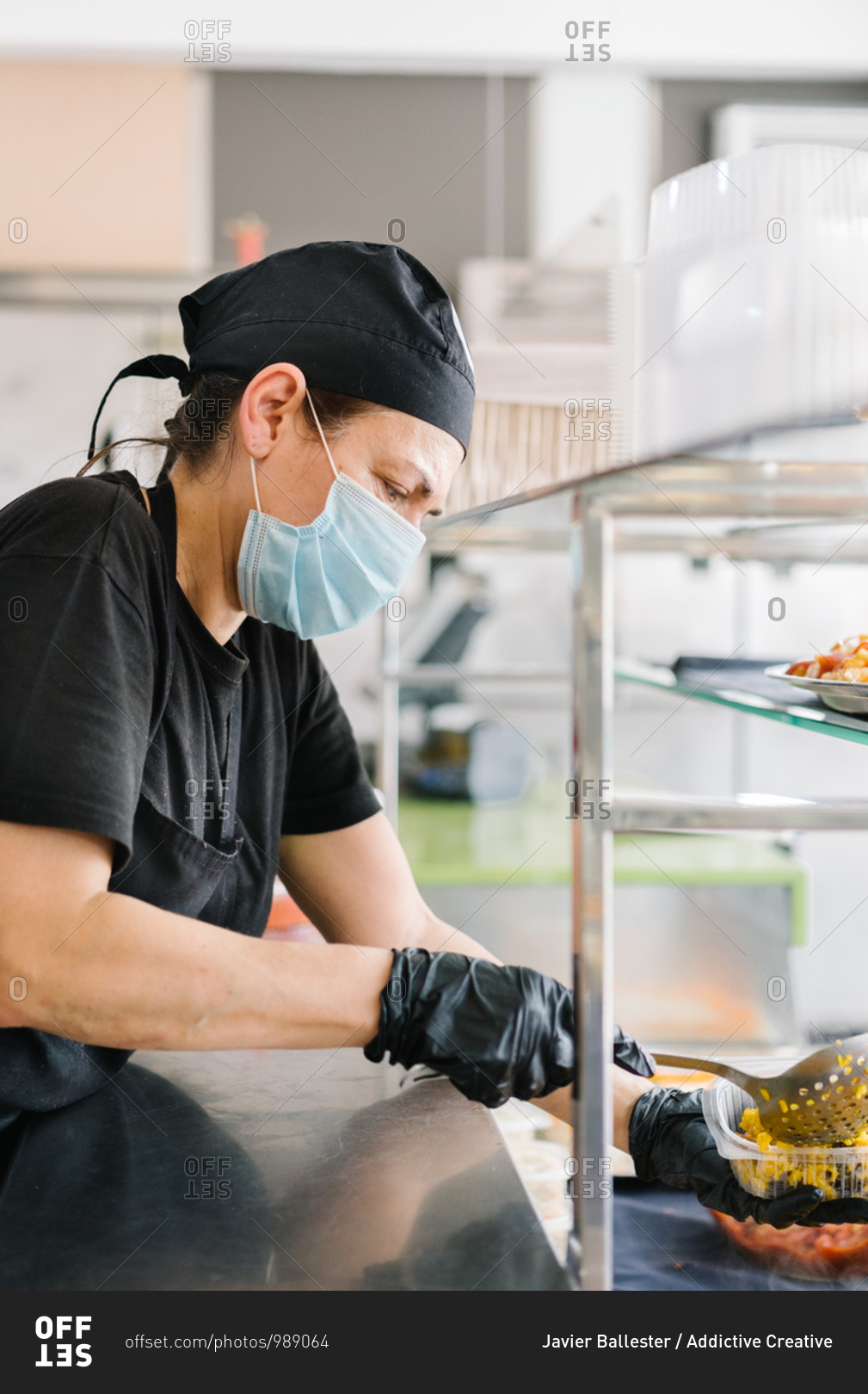 Side view focused waitress wearing face mask and gloves putting delicious dish into takeaway plastic container while working in cafe during coronavirus pandemic