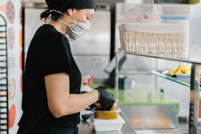 Side view o waitress in face mask putting fresh tasty dish into plastic takeaway container while working in cafe during coronavirus outbreak