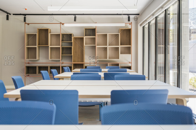 Modern style office interior with square tables and blue stools on floor and wooden shelves on wall with large window and lamps on ceiling