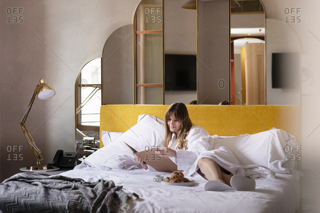 Young female in bathrobe lying on comfortable bed with cup of coffee and pastry and reading magazine while relaxing in cozy hotel room during holiday in Florence in Italy