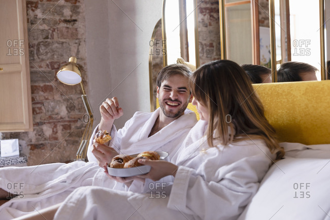 Side view of cheerful romantic couple in bathrobes sitting on bed and eating yummy pastry while enjoying breakfast in cozy hotel room with stylish interior in Italy