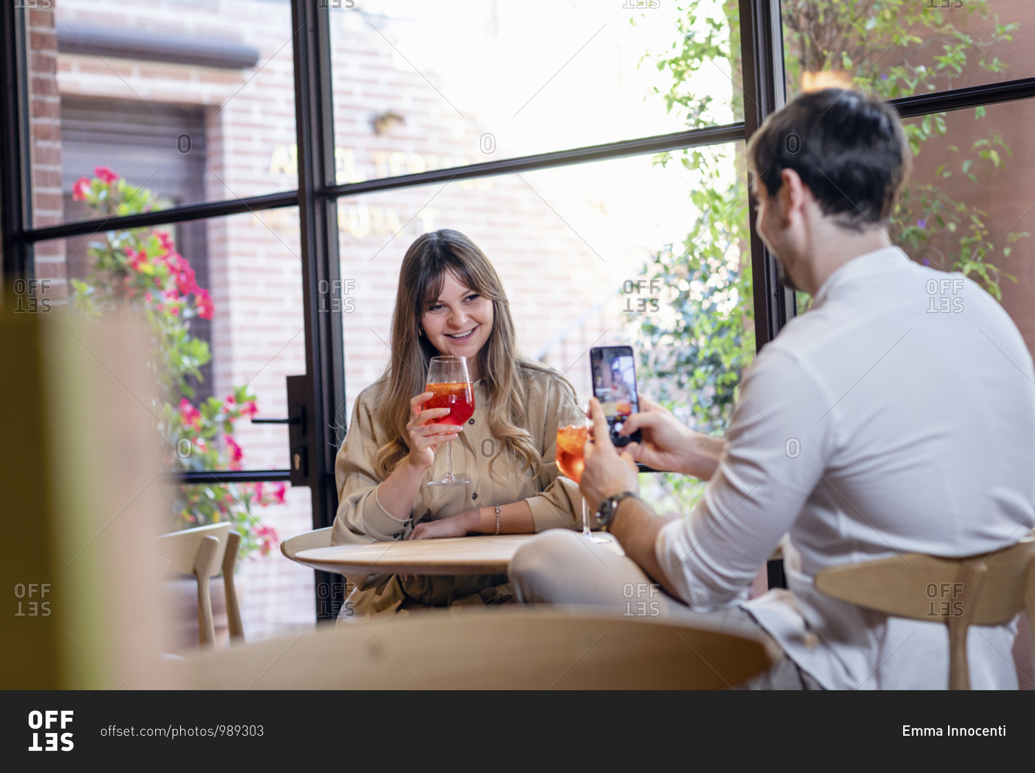 Young man with smartphone taking picture of happy girlfriend with glass of cocktail in hand while spending time together in cozy cafe