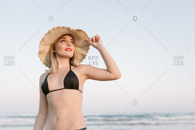 Young blonde woman with black bikini touching her hat at the beach while looking at infinity