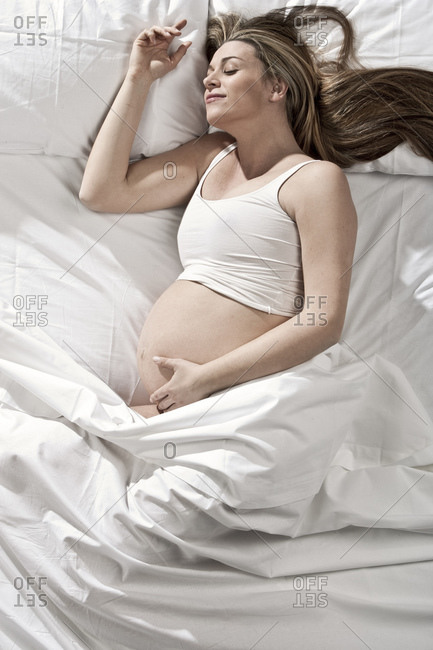 Portrait of heavily pregnant woman lying on bed, cradling stomach.