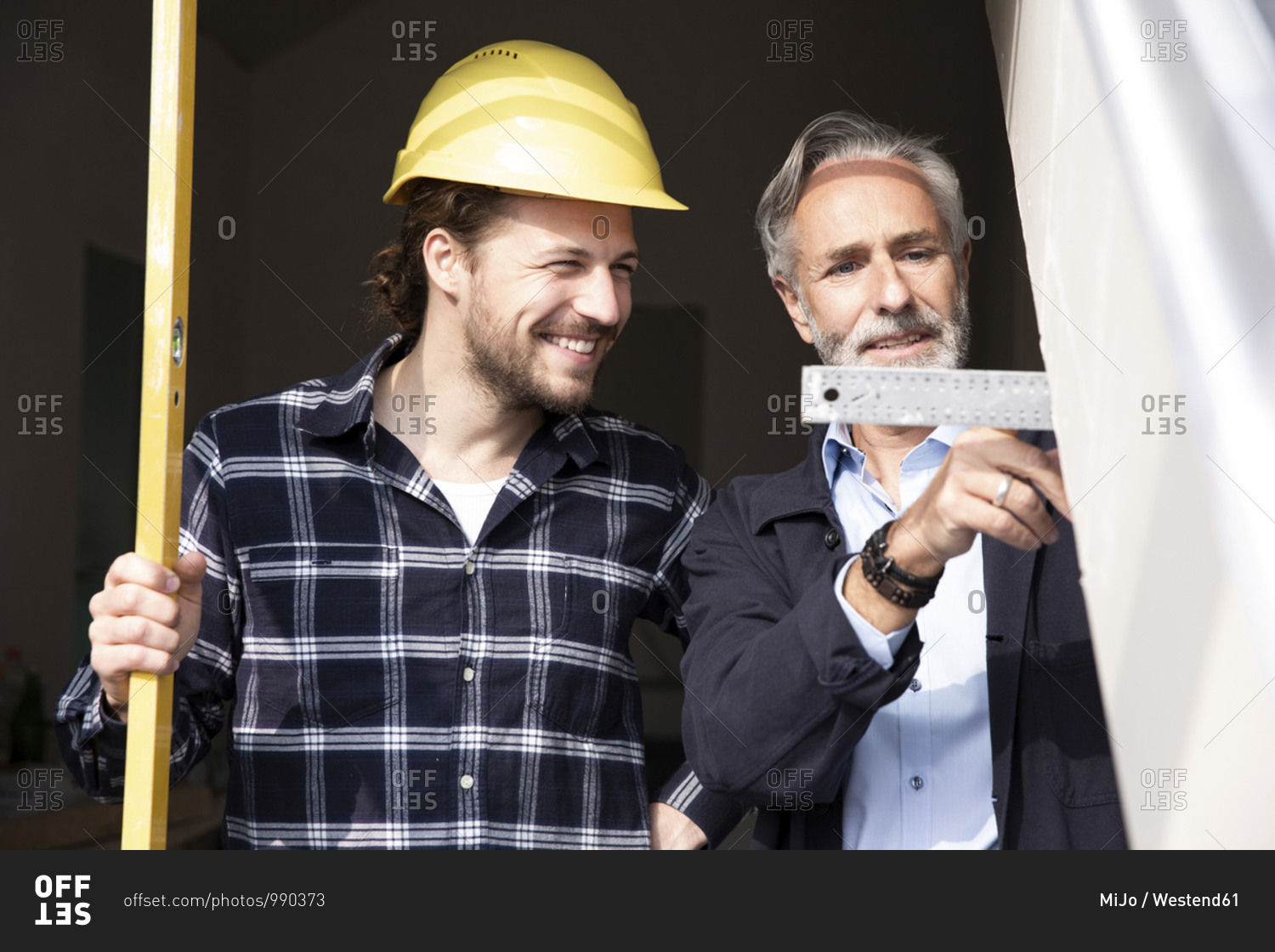 Smiling construction worker looking at measurement showing by architect in constructing house seen through window