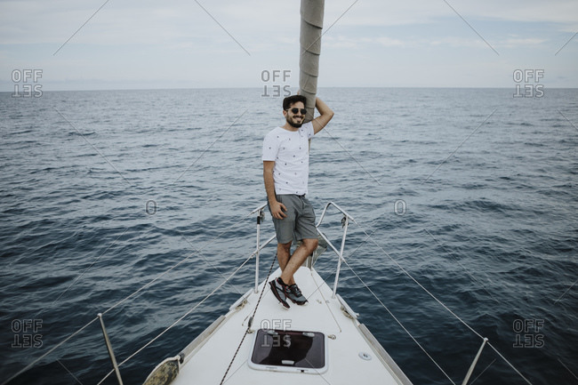 Male sailor wearing sunglasses standing on bow of sailboat in sea