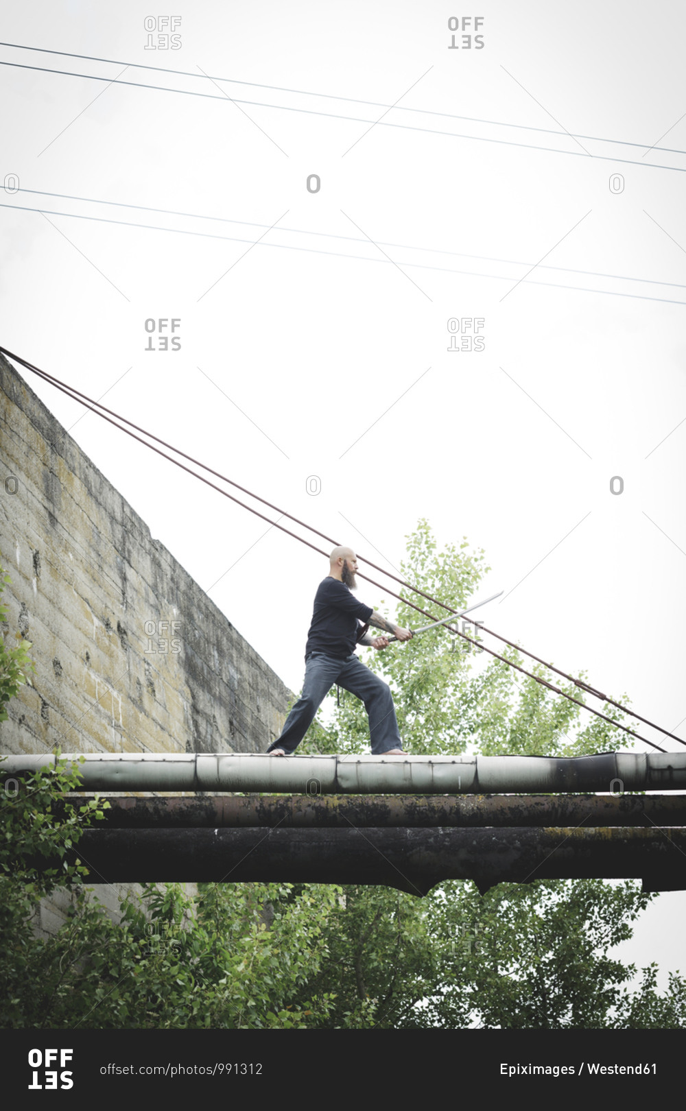 Mature man holding sword posing while standing on old pipes against clear sky