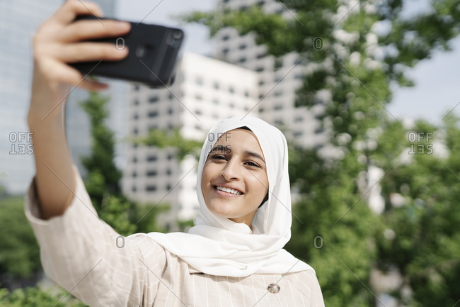Muslim teenage girl smiling while taking selfie through smart phone in city