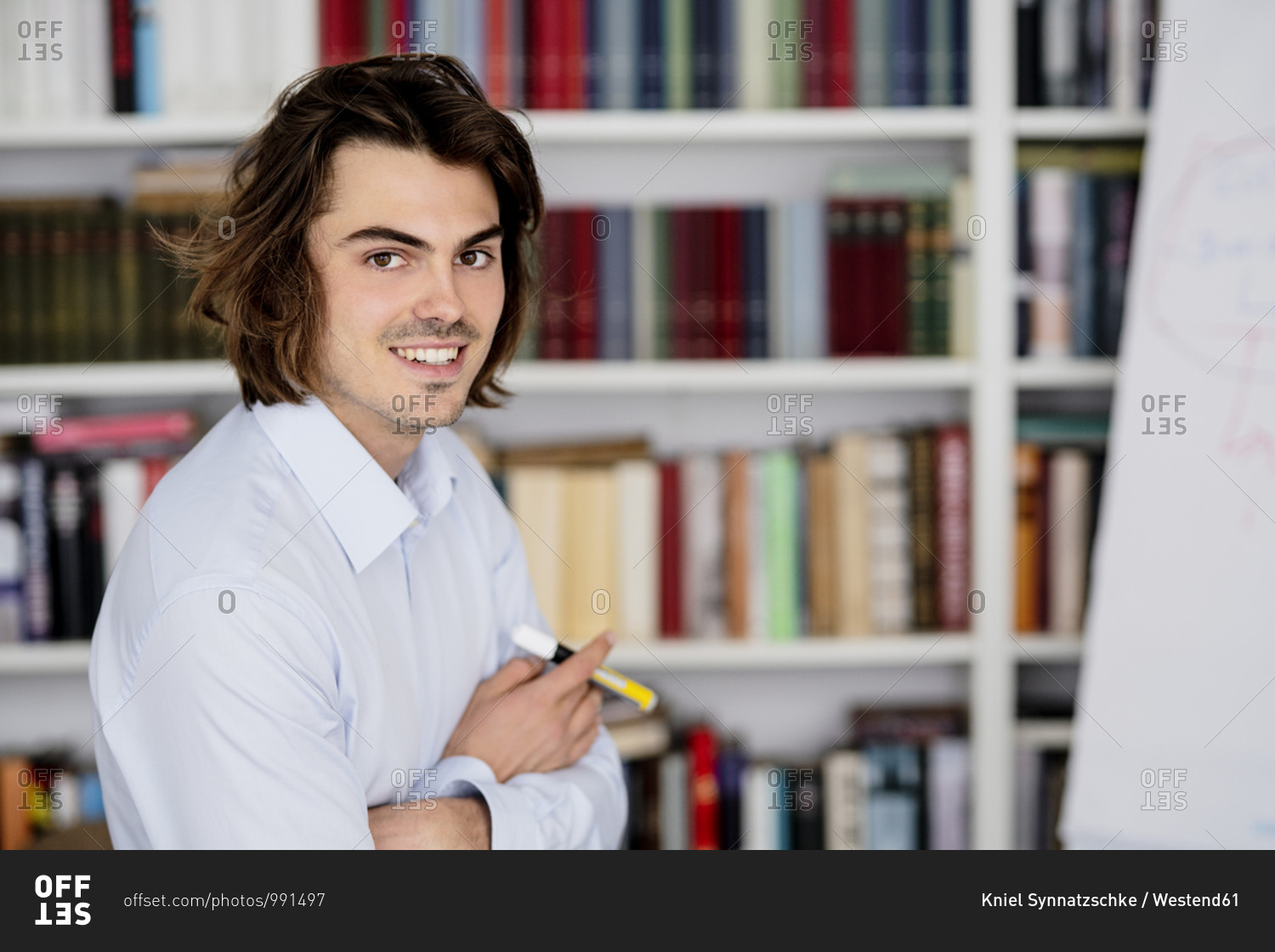 Smiling businessman with arms crossed sitting against bookshelf in office