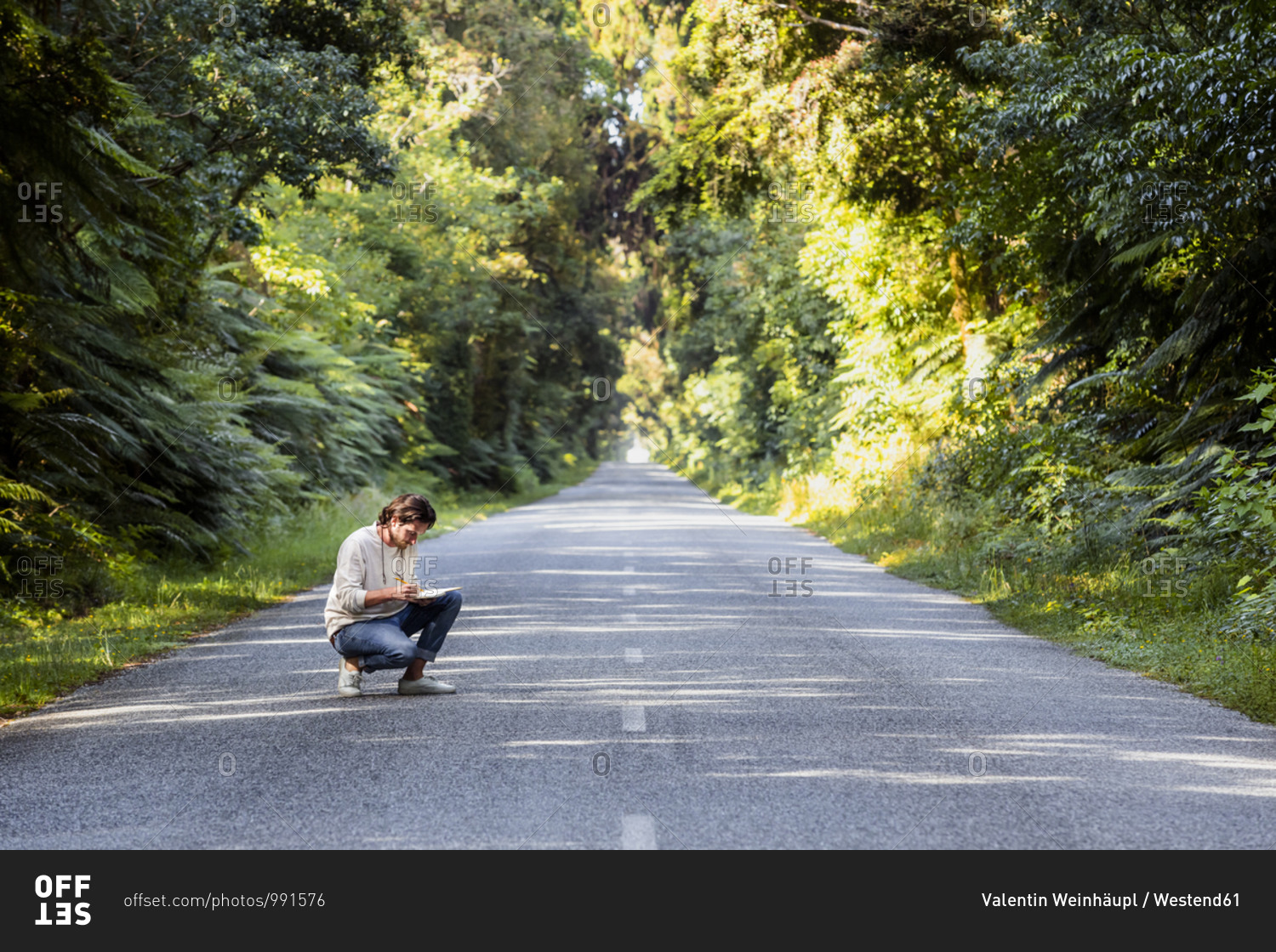 Young man writing in diary while crouching on country road amidst trees