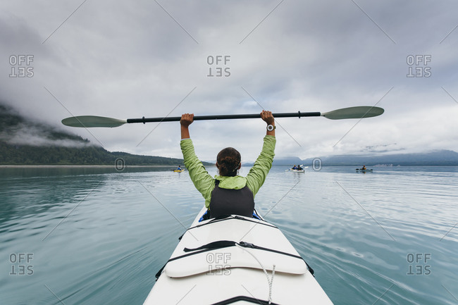 Sea kayaker extends paddle over her head, pristine waters of Muir Inlet in distance, Glacier Bay National Park, Alaska
