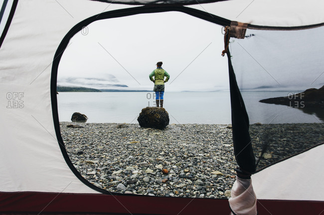 View through camping tent doorway of woman standing on beach, an inlet on the Alaska coastline.