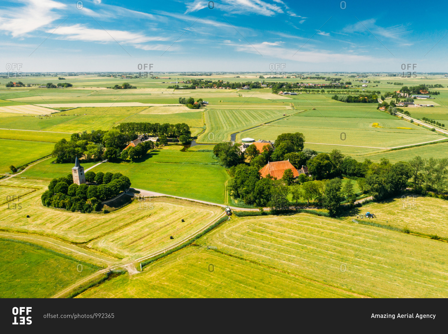 Aerial view of tractor mowing the grass on farmland with farmer houses and a little church in Friesland, The Netherlands.