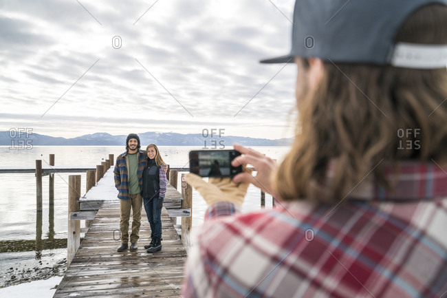 Friend takes a cellphone picture for a couple standing at the end of a pier