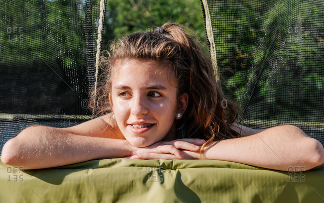 Horizontal portrait of a curly brunette teenager very happy smiling while lying on a trampoline with the net at the background. The girl feels happy and tired after jumping on the trampoline.