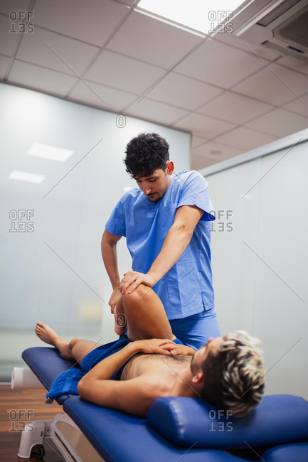 Osteopath in blue uniform examining leg of unrecognizable slim male patient with dyed hair lying on examination table in clinic
