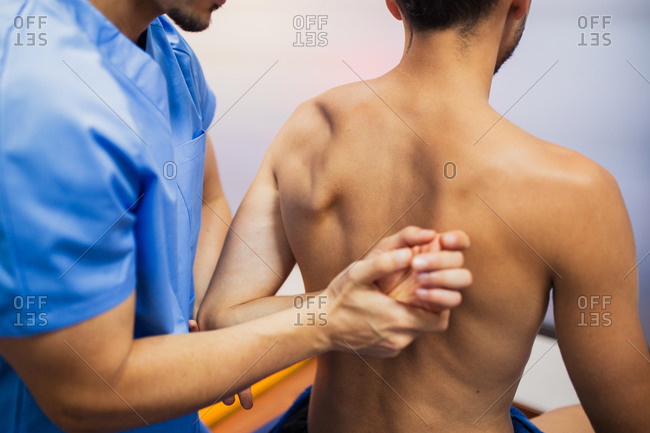 Crop of back view of orthopedist in uniform checking up shoulder joint of fit man sitting on examination couch with flexed arm in hospital and looking away