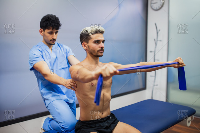 Sports medicine doctor checking up back of sportive man with dyed hair stretching elastic tape during rehabilitation period and looking away