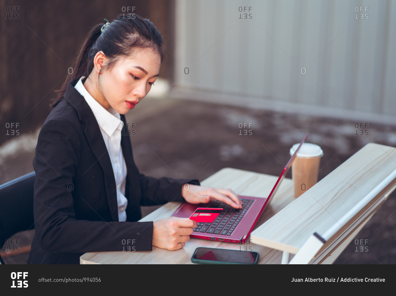 Side view of serious young Asian business lady in formal suit with credit card using laptop and smartphone during online payment while sitting at table at workplace on terrace