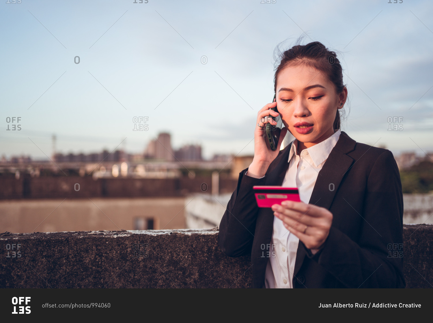Serious young Asian businesswoman in formal wear holding credit card and having phone conversation with bank customer service while standing on rooftop of city building