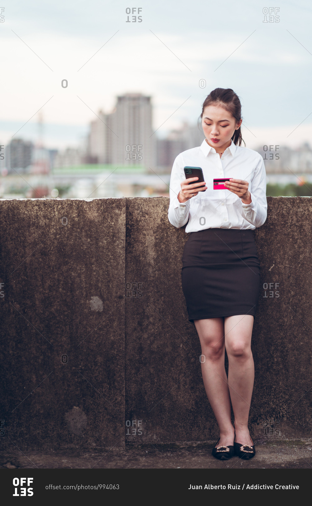 Serious young Asian businesswoman in formal wear holding credit card and smartphone with bank customer service while standing on rooftop of city building
