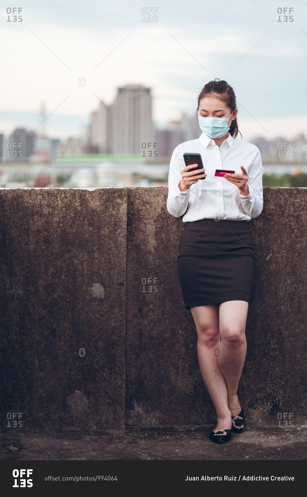 Young Asian female remote employee in formal clothes and medical mask holding credit card and using mobile phone during online payment while standing on rooftop of modern city building