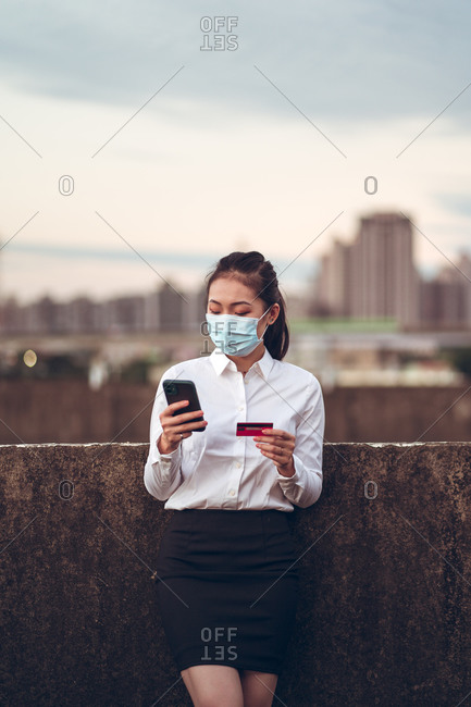 Young Asian female remote employee in formal clothes and medical mask holding credit card and using mobile phone during online payment while standing on rooftop of modern city building