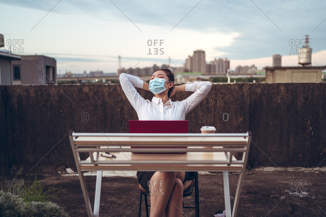 Young Asian female employee in formal wear and protective mask sitting at table with laptop and cup of coffee on rooftop terrace and relaxing after remote work during coronavirus pandemic