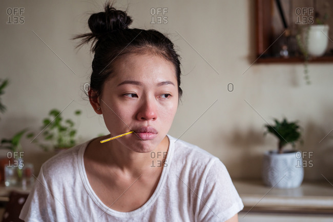 Exhausted ethnic female sitting on bed at home and measuring temperature with thermometer while having cold and looking away