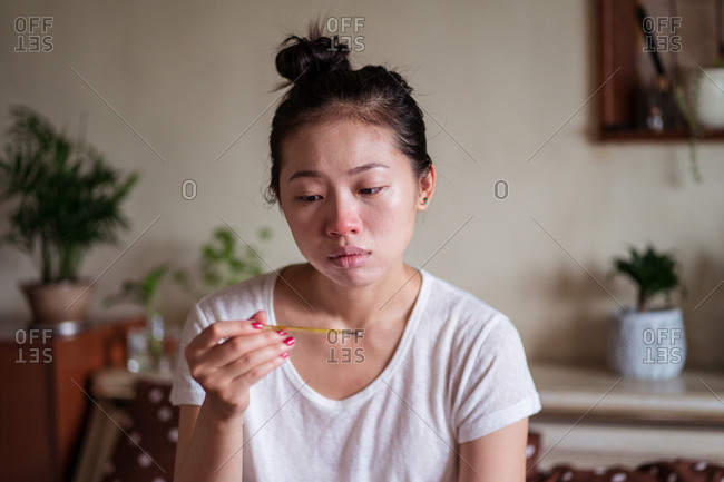 Exhausted ethnic female sitting on bed at home and measuring temperature with thermometer while having cold and looking away