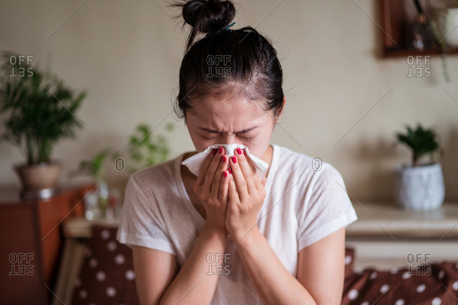 Frowned ethnic female in domestic clothes sitting on bed with running nose while having cold