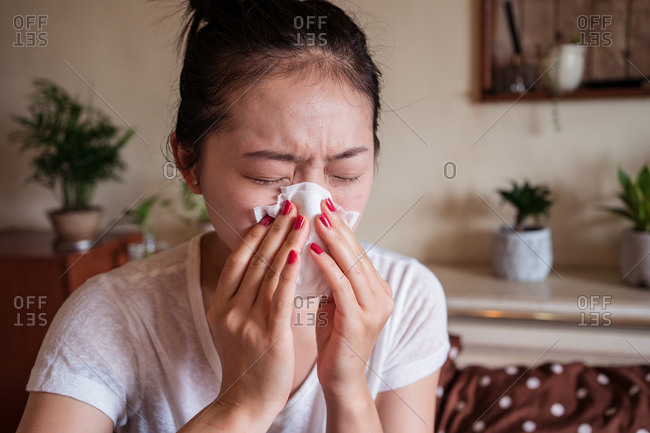 Frowned ethnic female in domestic clothes sitting on bed with running nose while having cold
