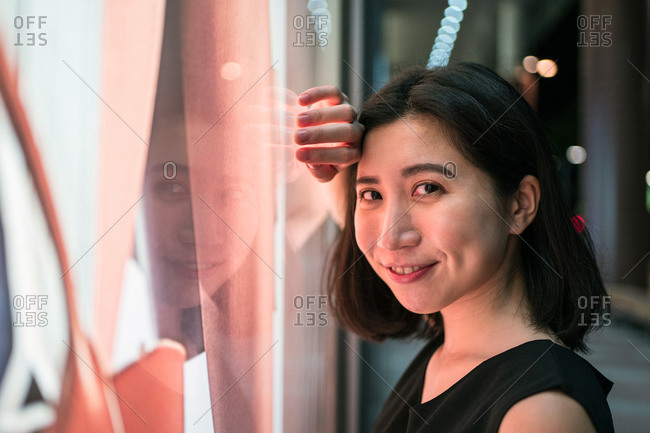 Content Asian female leaning on window illuminated by neon lights and looking at camera
