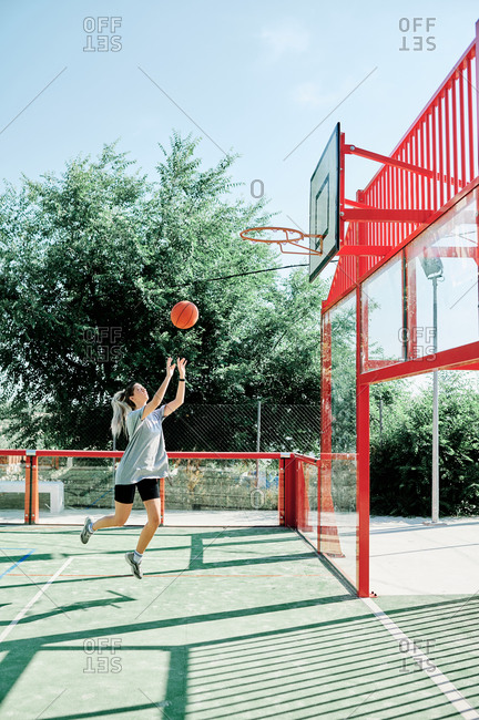 Full body side view of young sporty woman performing jump shot while playing basketball alone on sports ground