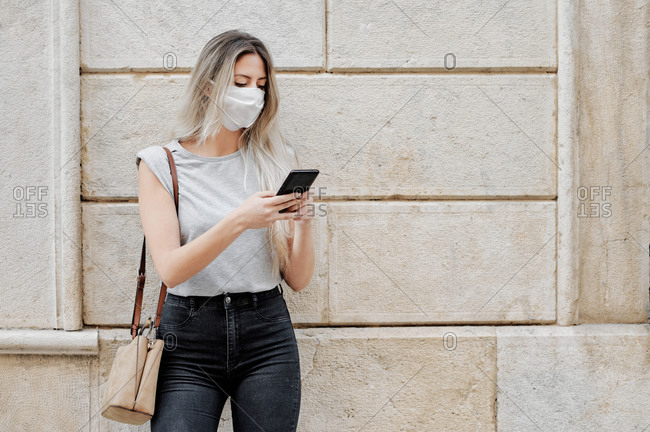 Young female in stylish outfit wearing a face medical mask messaging on mobile phone while standing against old stone concrete building city