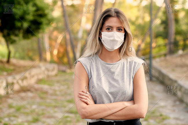 Young female in casual clothes and protective mask for coronavirus prevention looking at camera while standing on alley in green park