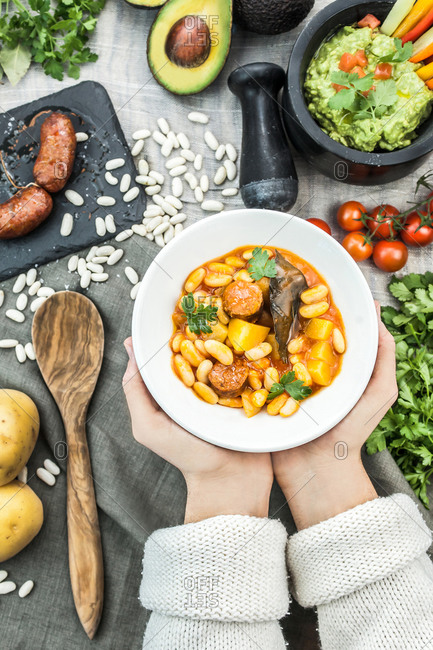 Top view of faceless person with bowl of white bean soup with chopped pork sausages and vegetables above table with ingredients and guacamole in mortar